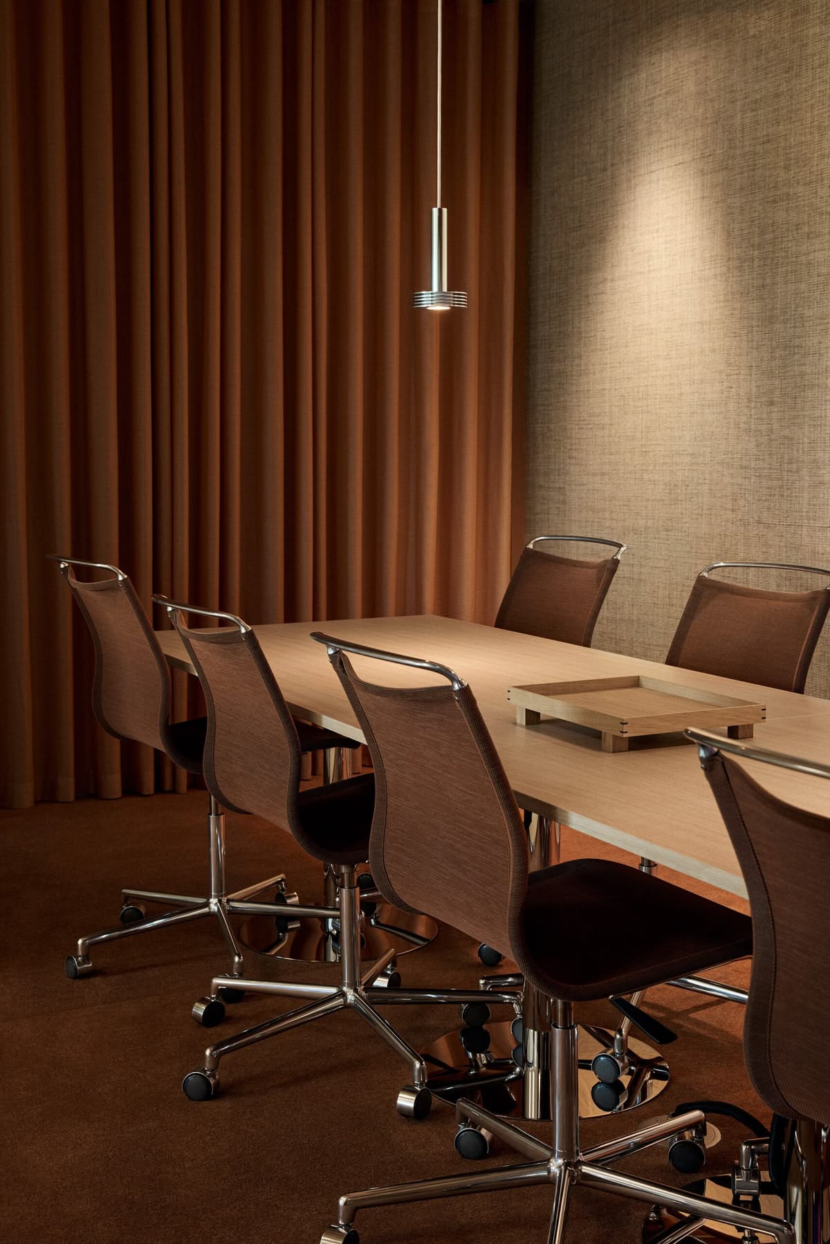 Meeting room with a pale timber table, brown mesh chairs and a pendant light against rich terracotta curtains — purposeful and refined