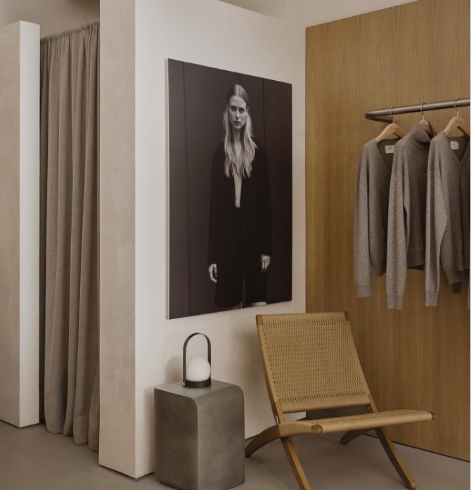 Minimal fashion retail interior with large-format portrait artwork, rattan chair and clothing rail — restrained and editorial