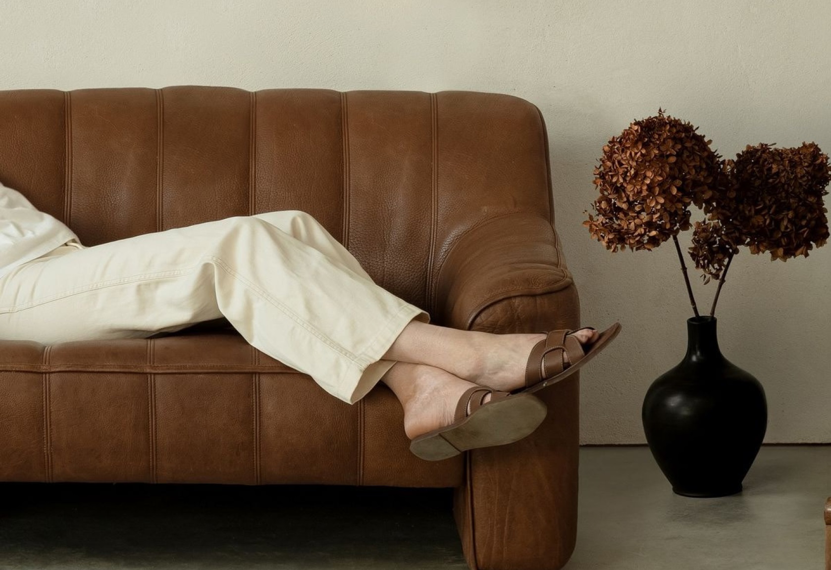 A person resting on a richly textured brown leather sofa beside a dark vase of dried flowers — warmth, material richness and considered atmosphere